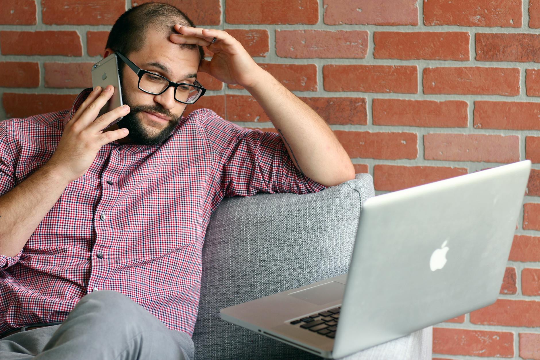 Zrezygnowany mężczyzna z brodą patrzy w laptop, trzymając telefon